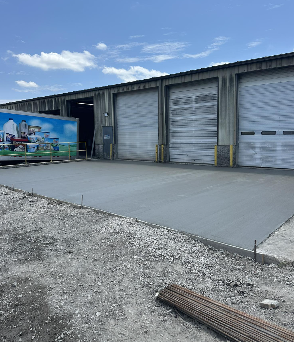 Newly poured concrete floor in front of a warehouse with three large metal garage doors, one partially open, and a vending truck with dairy products parked nearby. The area is outdoors with a gravel surface and construction materials in the foregroun