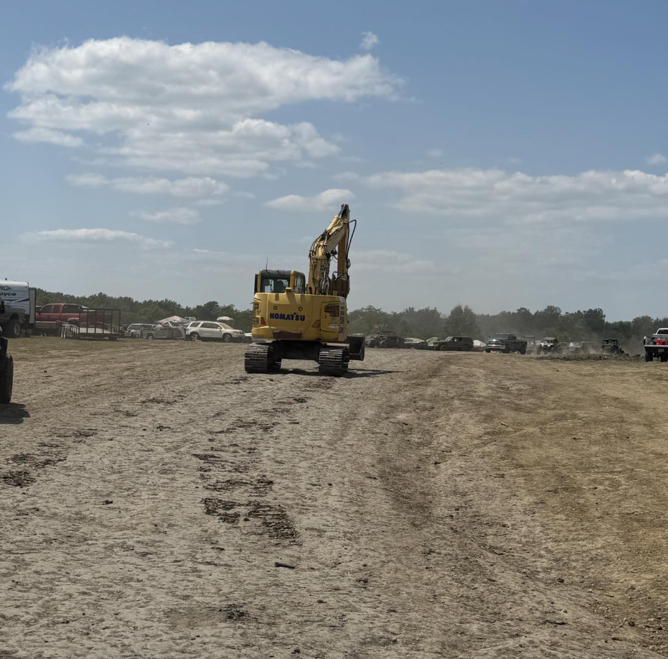 A yellow Komatsu excavator working on a dirt area with a line of parked cars and trucks in the background under a partly cloudy sky.