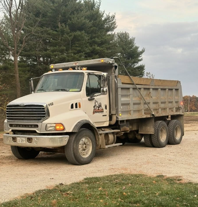 A white Sterling dump truck parked on a dirt surface with trees in the background.