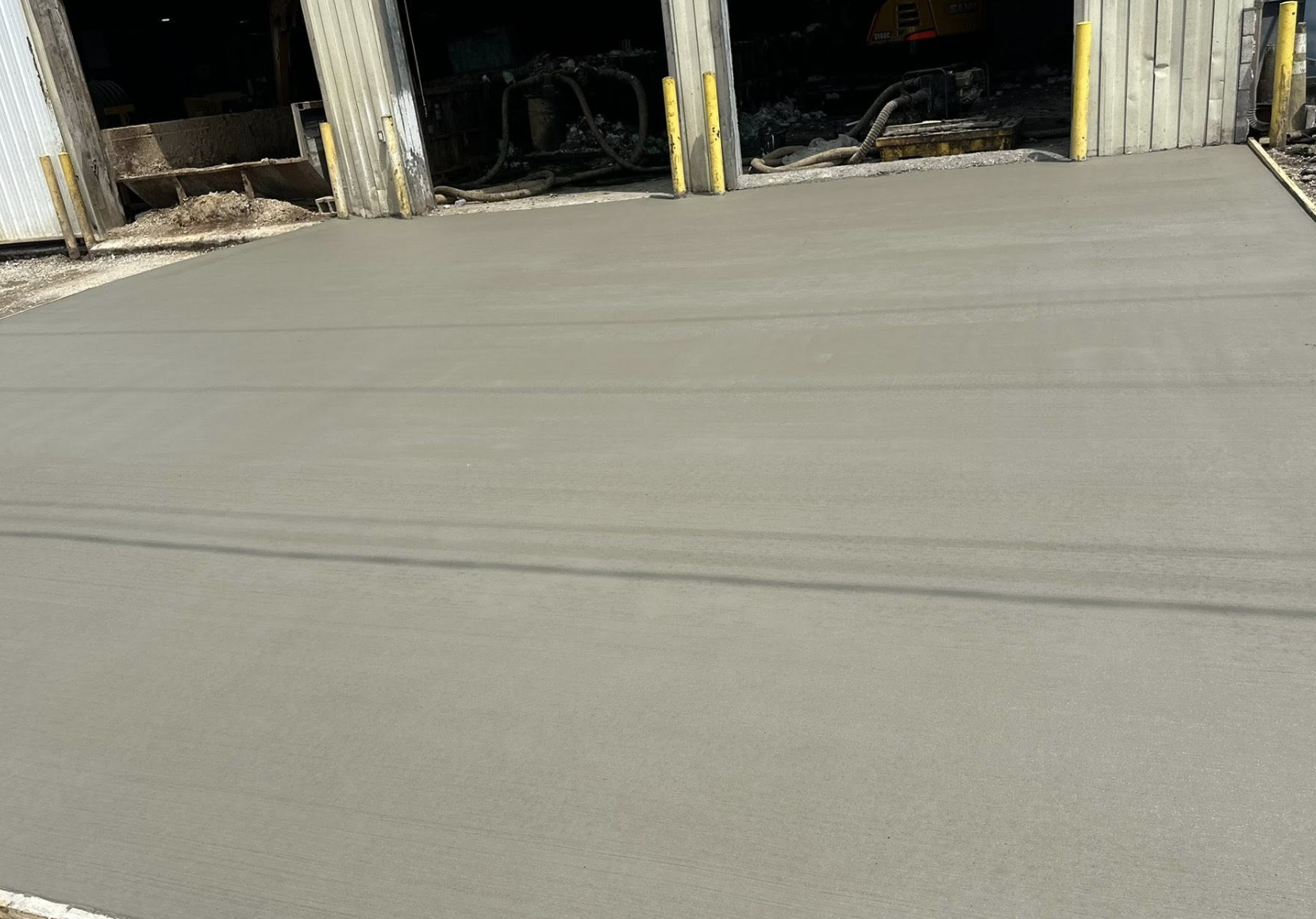 Freshly poured concrete slab outside an industrial building with partially open large metal doors, yellow safety posts, and construction equipment inside.