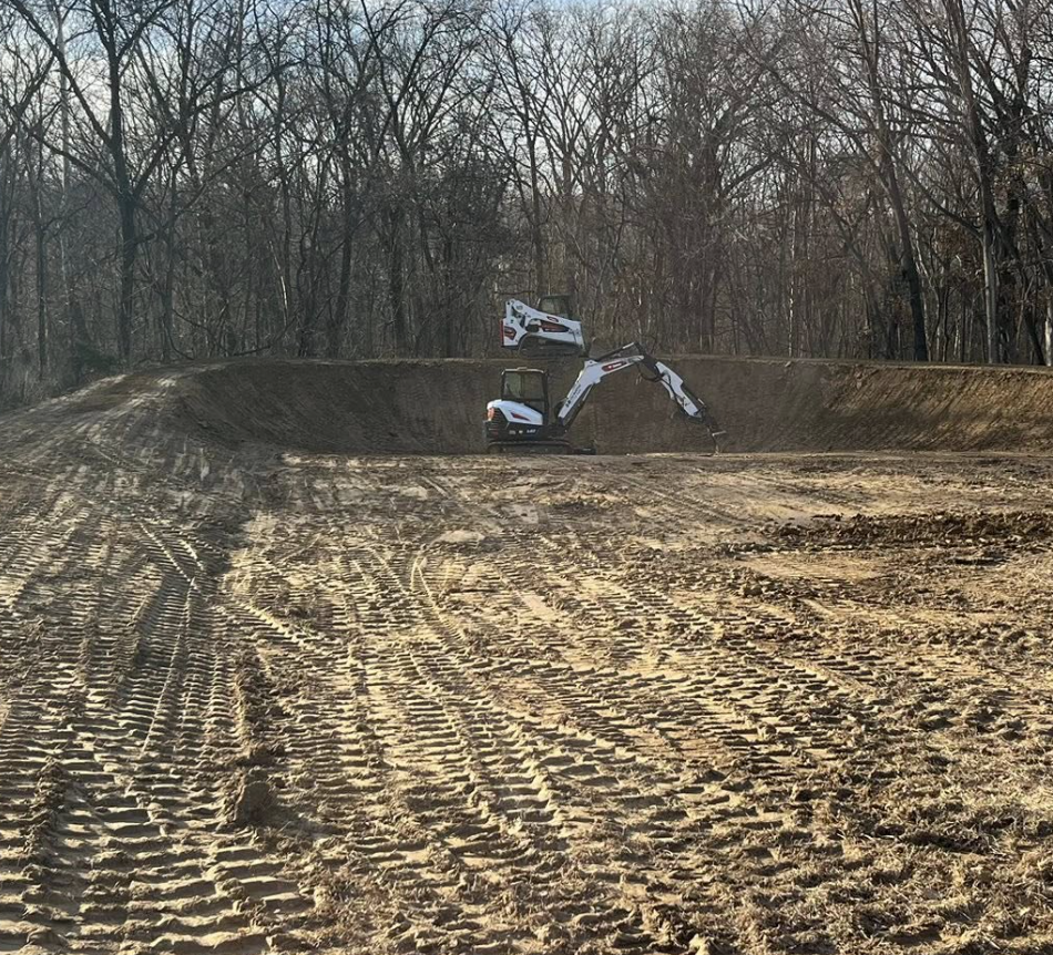 Construction site with toy construction vehicles, including a mini excavator and two trucks, in a cleared dirt area surrounded by leafless trees.