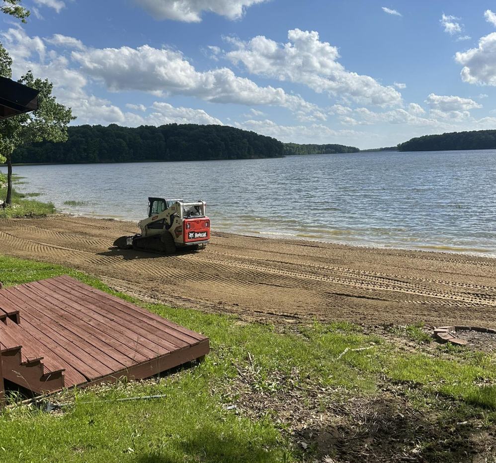 Lakeside scene with a small Bobcat construction vehicle on a sandy beach, a wooden deck on green grass, calm water, trees, and a partly cloudy sky.