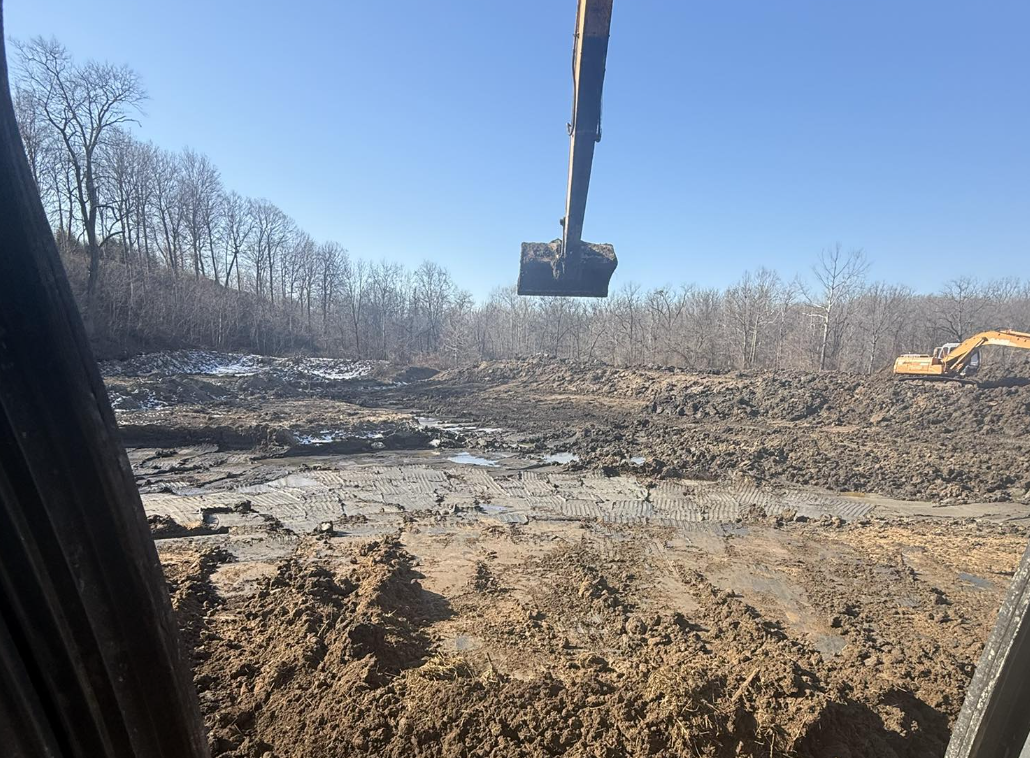 Construction site with excavator and muddy terrain under a clear blue sky, trees in the background.