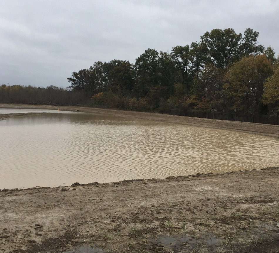 A muddy, low water level pond or reservoir with a fence on the far side and trees with fall foliage in the background.