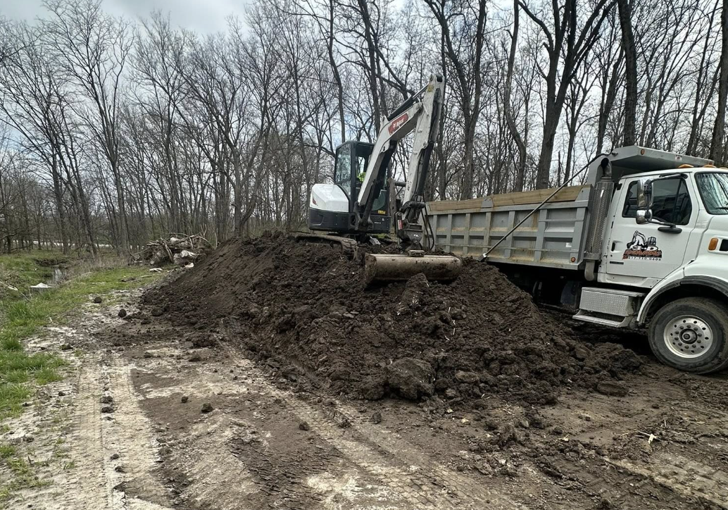 A small excavator on top of a dirt pile next to a dump truck, with trees in the background, on a dirt road.