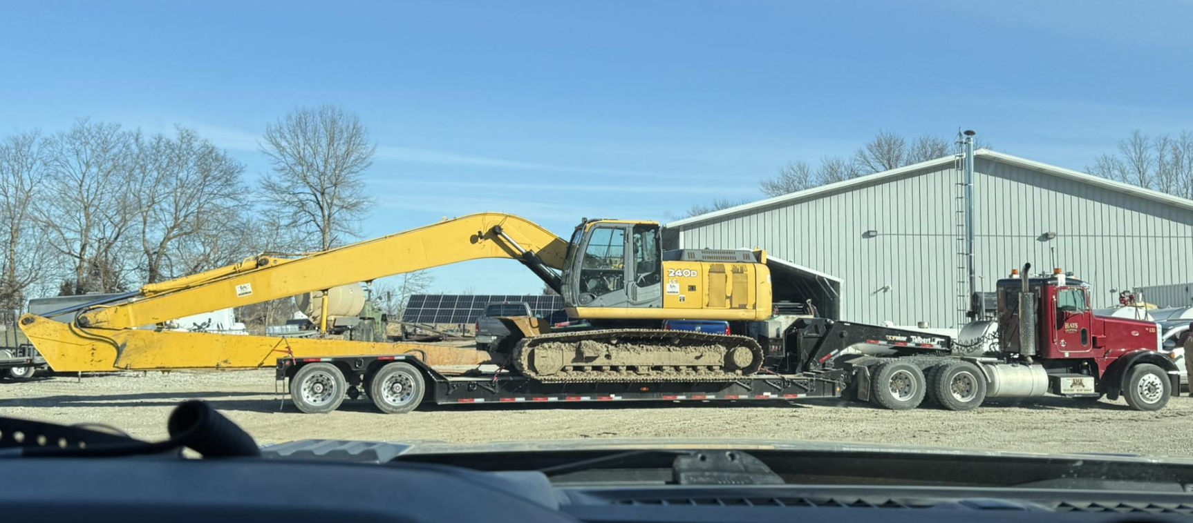 A yellow excavator on a flatbed trailer attached to a red semi-truck, with a warehouse and trees in the background.