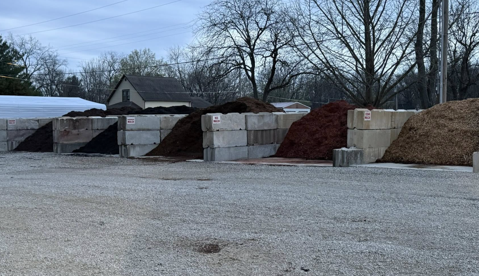 Several large piles of different types of mulch behind concrete block walls outdoors, with trees, a house, and utility wires in the background.