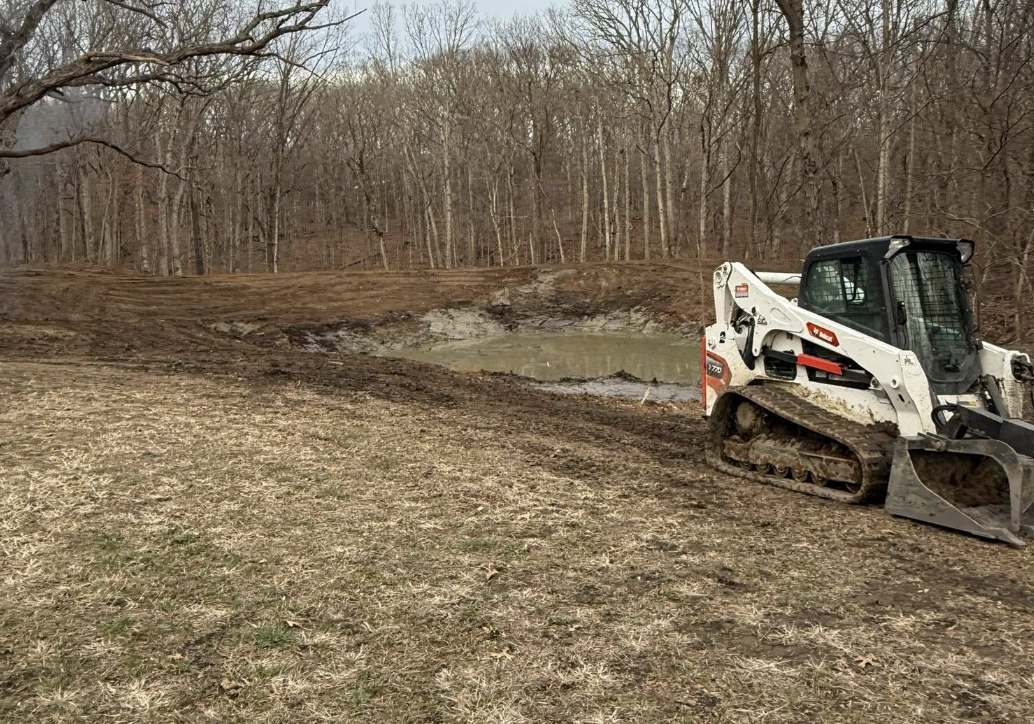 A small excavator parked near a muddy pond in a wooded area, with leafless trees surrounding the scene.