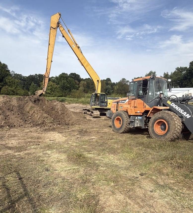 Construction site with an orange Doosan wheel loader and a yellow excavator digging soil amidst a grassy area with trees in the background.