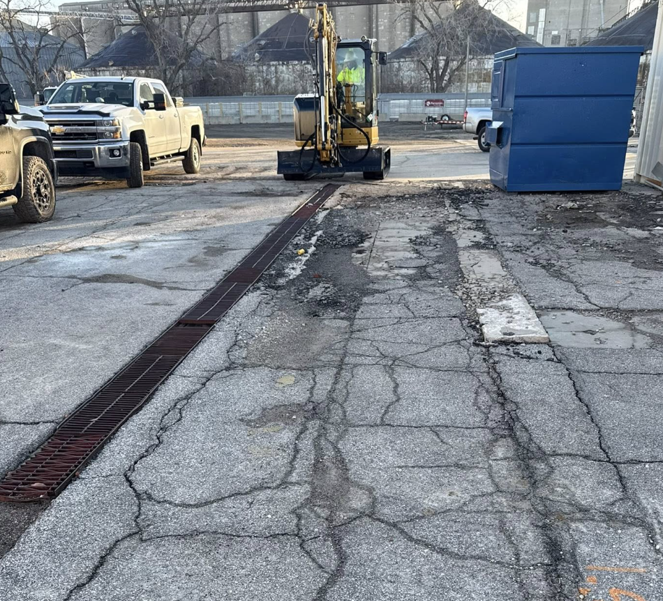 View of a parking lot with cracked asphalt, a drainage grate, a small excavator, and parked trucks. Construction work appears to be ongoing.