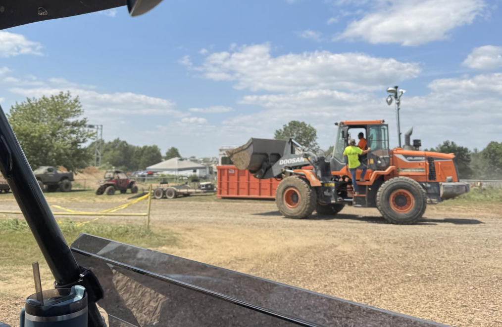 An orange front loader carrying dirt or gravel at a construction site with workers and parked vehicles in the background under a blue sky with clouds.