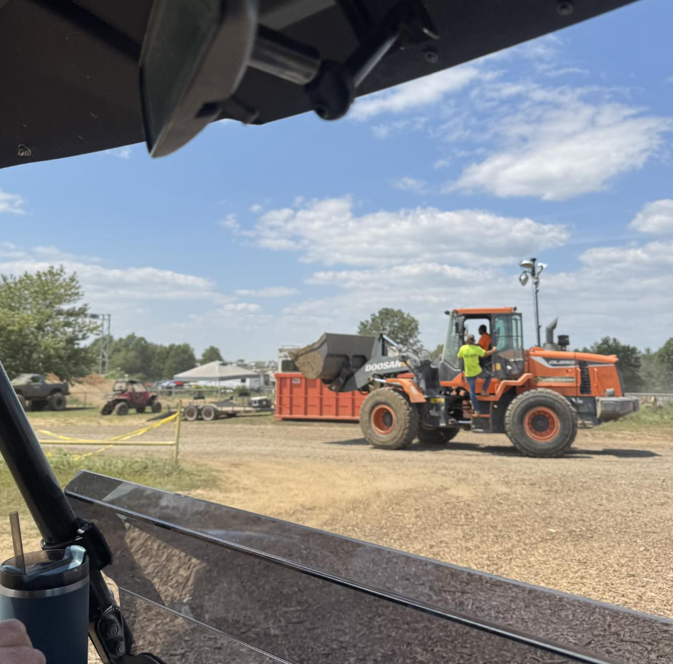 View from inside a vehicle looking out at a construction site with a large orange front loader moving dirt into a red container, with workers nearby and a partly cloudy sky.