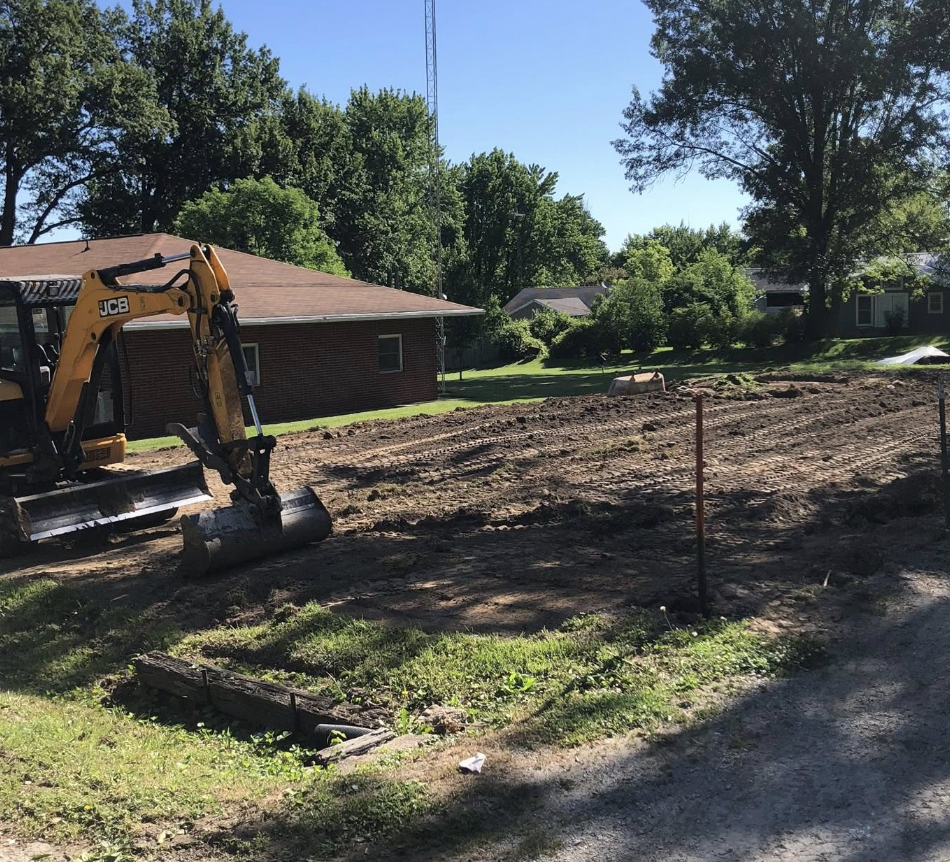 Construction site with a small excavator working on leveling the ground in a residential area with trees and houses in the background.