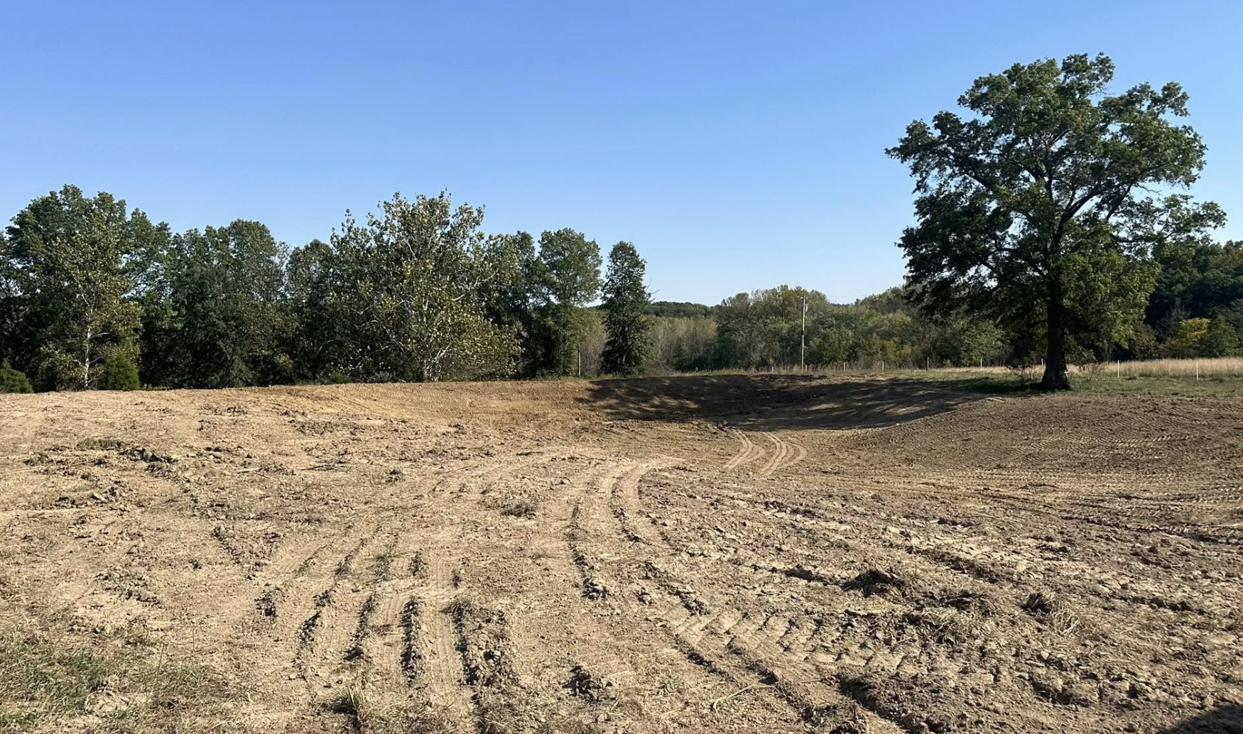 A cleared dirt field with visible tire tracks, a large tree on the right, and a line of trees in the background under a clear blue sky.