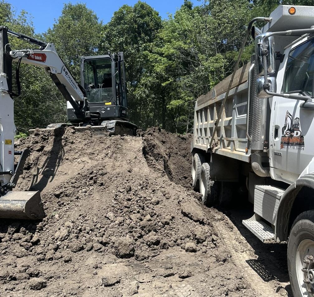 A construction site with a small excavator loading dirt into a dump truck surrounded by trees.