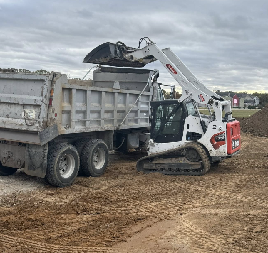Small excavator loading dirt into a dump truck on a construction site with cloudy skies.