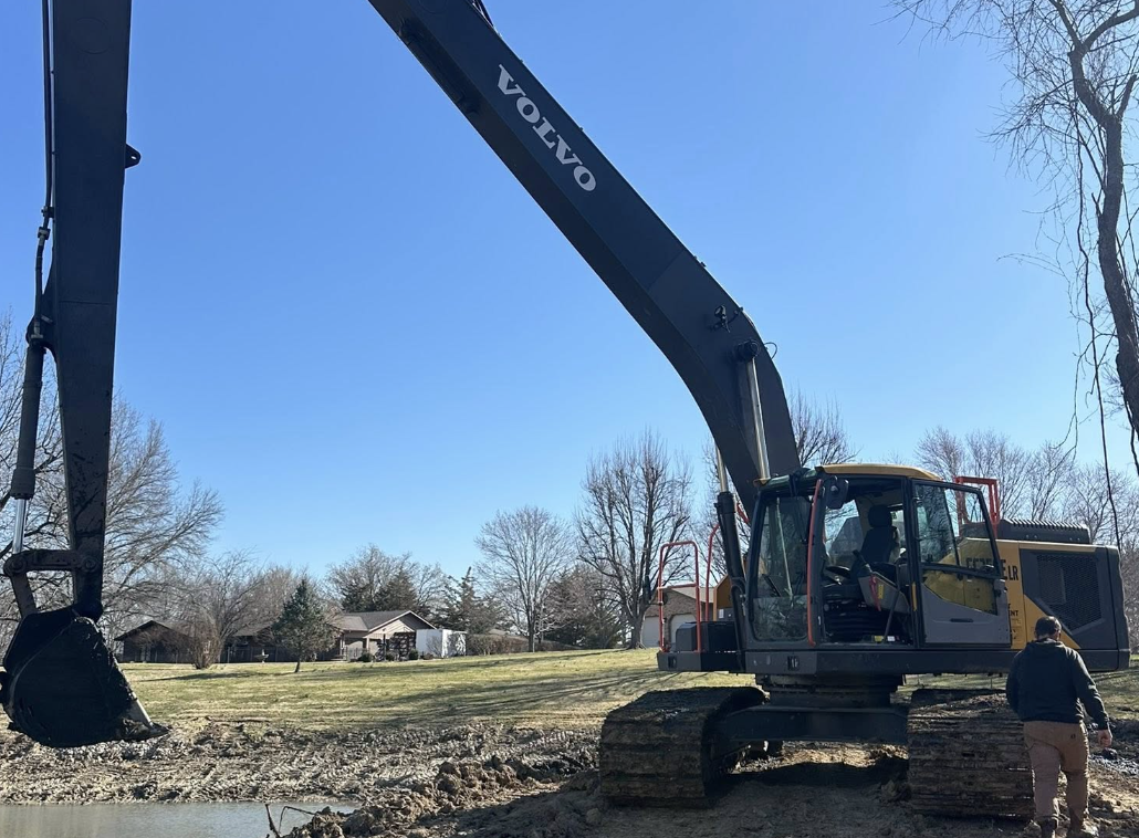 A large Volvo excavator operating on a construction site next to a small body of water, with a person standing nearby and houses visible in the background.