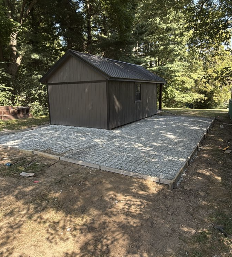 Small brown shed on a newly paved concrete patio in a wooded backyard.