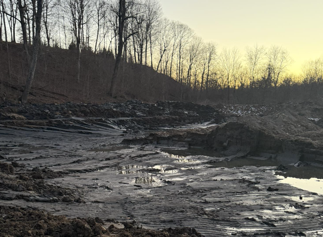 A muddy, partially dried riverbed with water puddles and leafless trees in the background during sunset.
