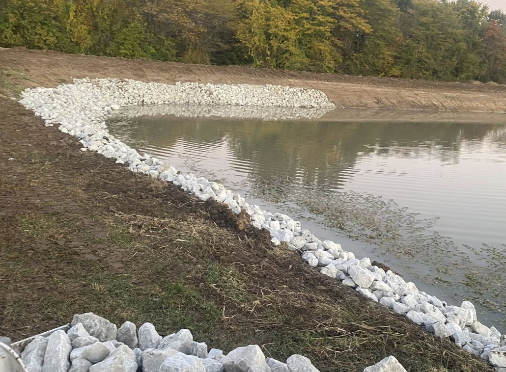A lake or pond with a shoreline reinforced by white rocks, with trees in the background.
