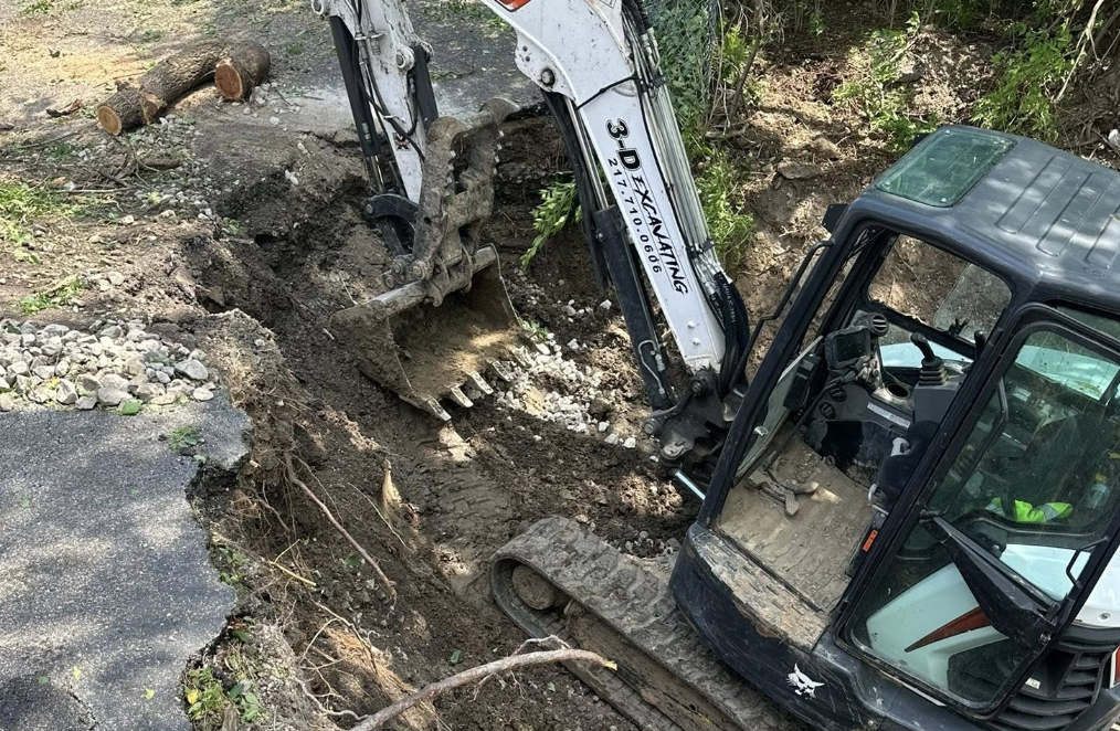 A backhoe excavator digging a trench on a dirt surface near a paved area and some logs.