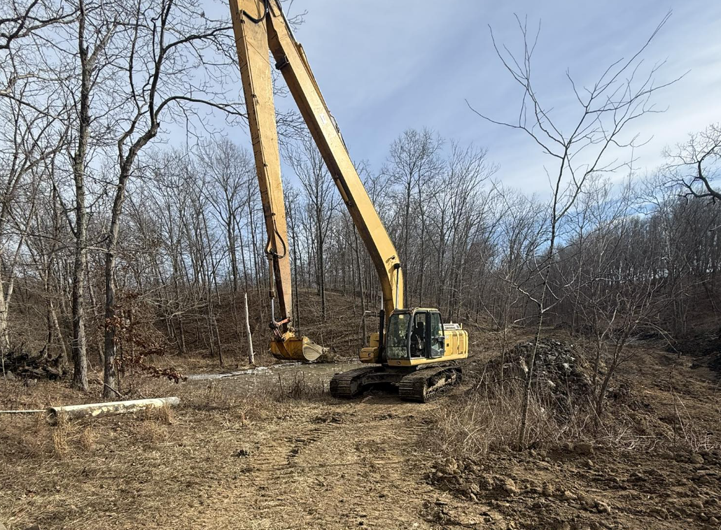 A large yellow excavator working in a wooded area with leafless trees, digging into the dirt on a clear day.