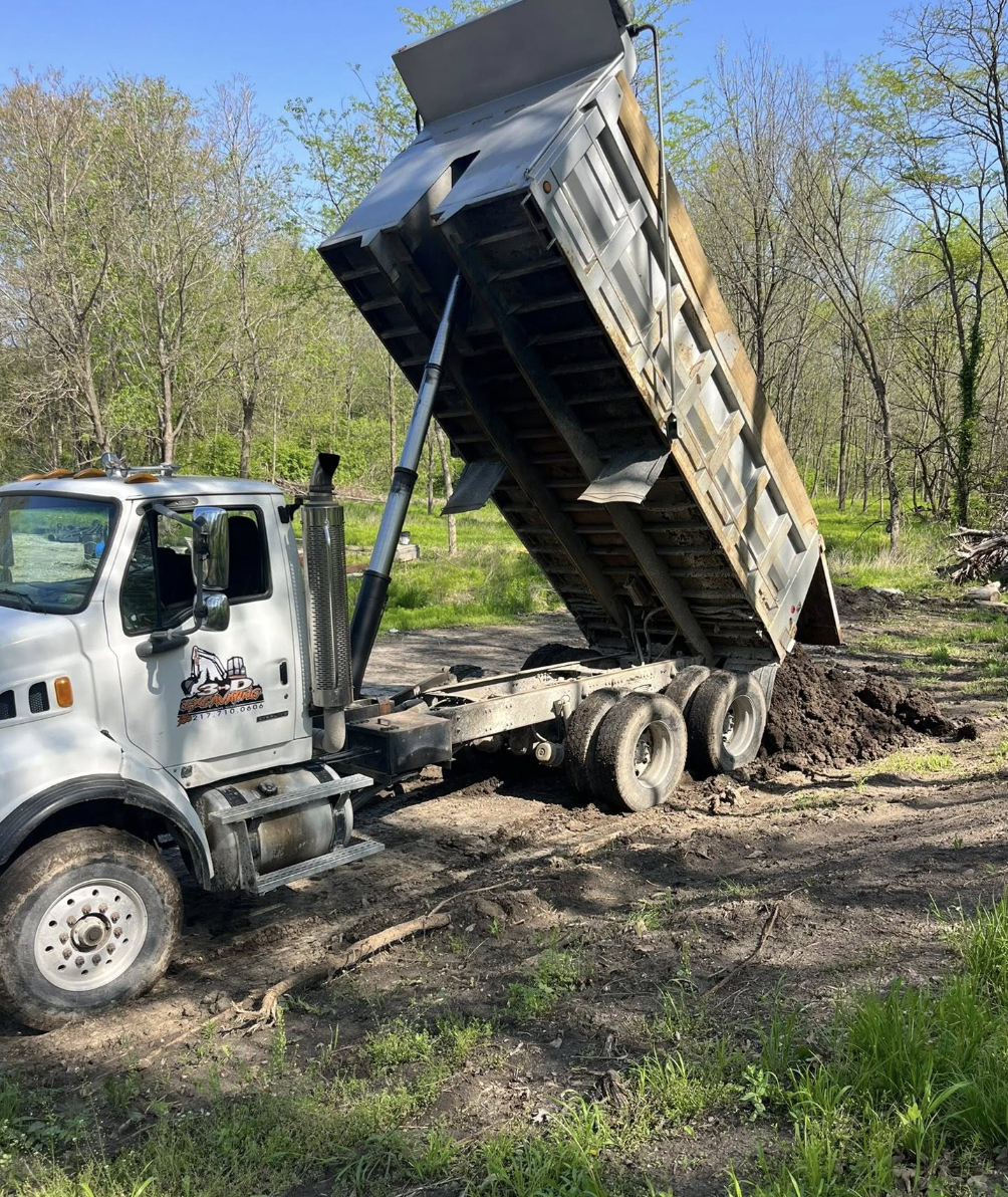 A dump truck unloading dirt on a grassy outdoor construction site with trees and blue sky in the background.