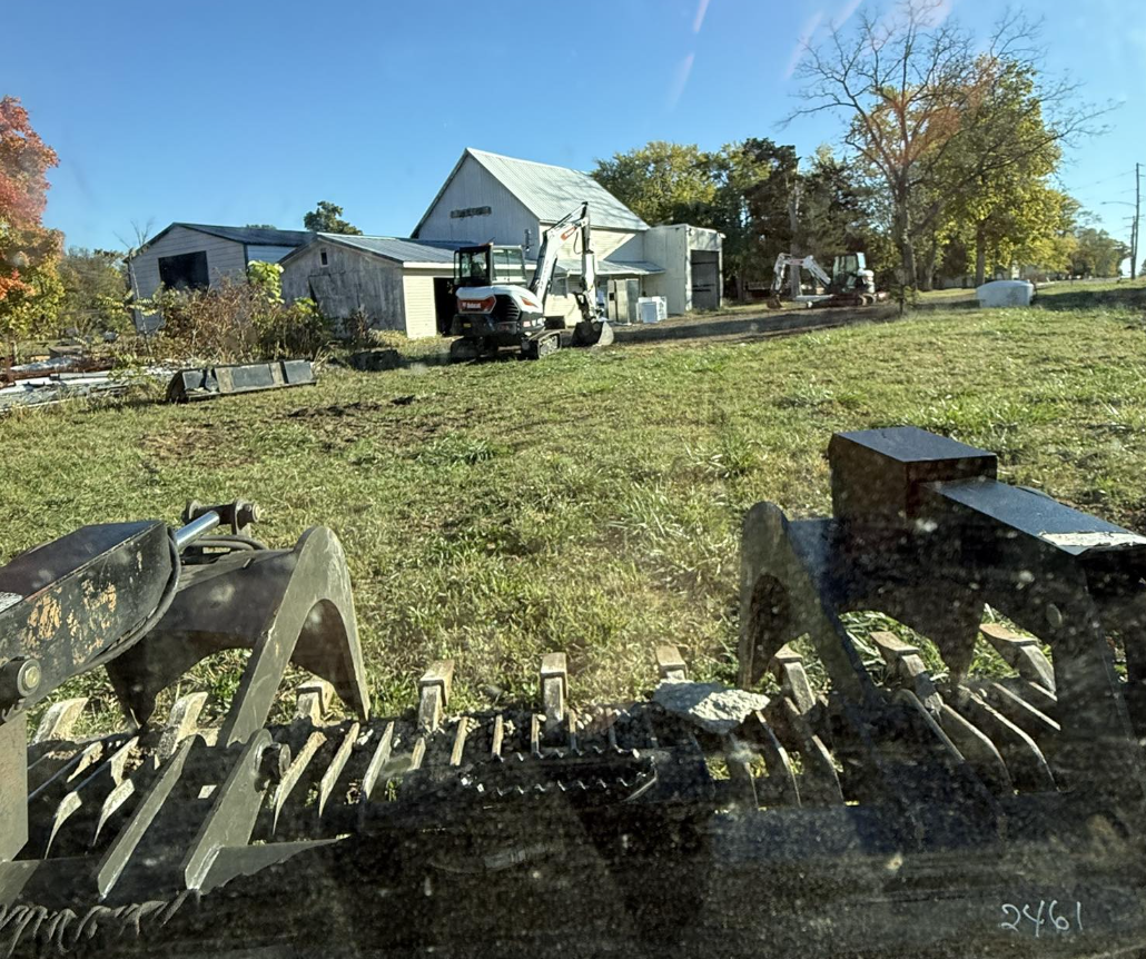 View of a farm with multiple small buildings and construction equipment, including a mini excavator and another piece of machinery, on a grassy plot of land during daytime with clear blue skies and trees in the background.