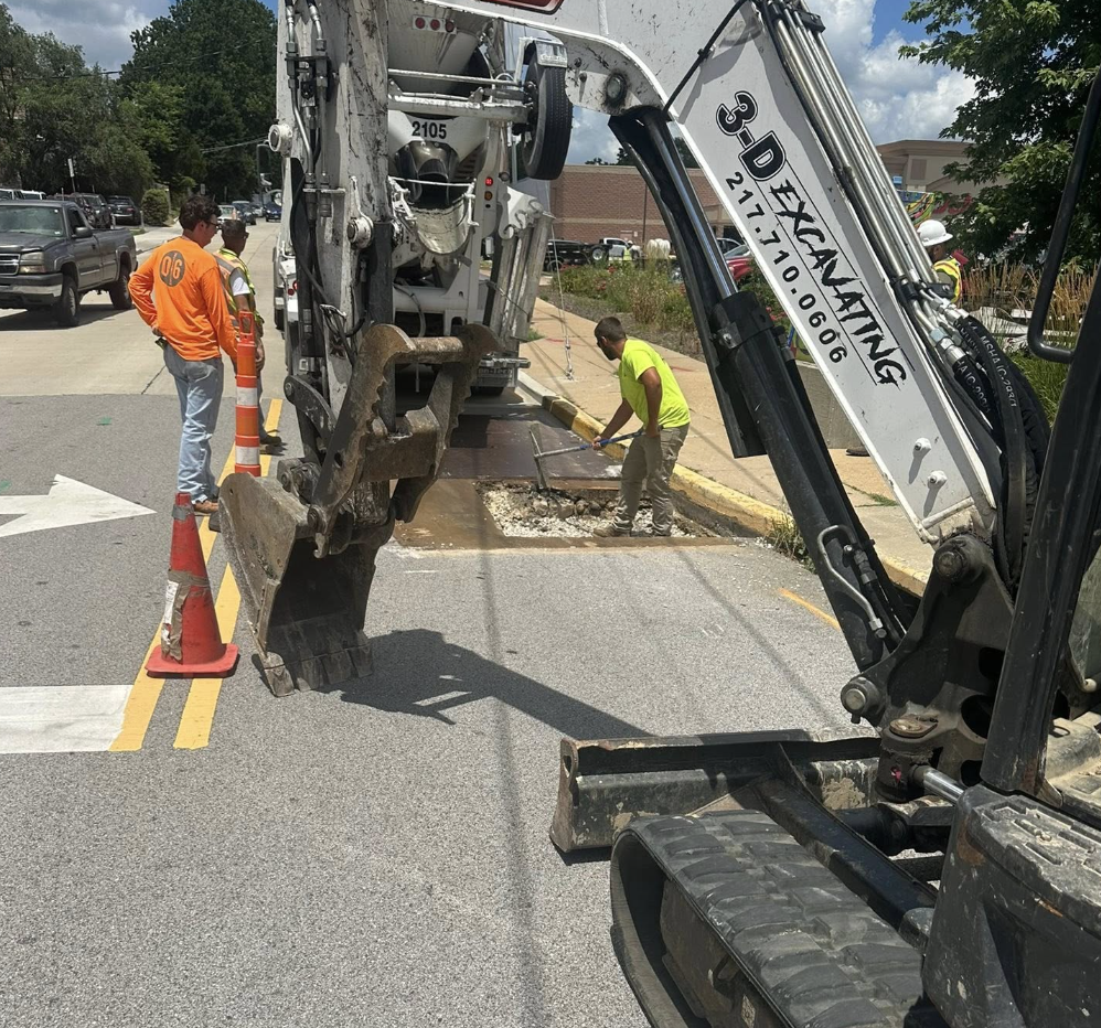 Construction workers and a backhoe digging up a section of asphalt on a city street, with cars parked and driving nearby.