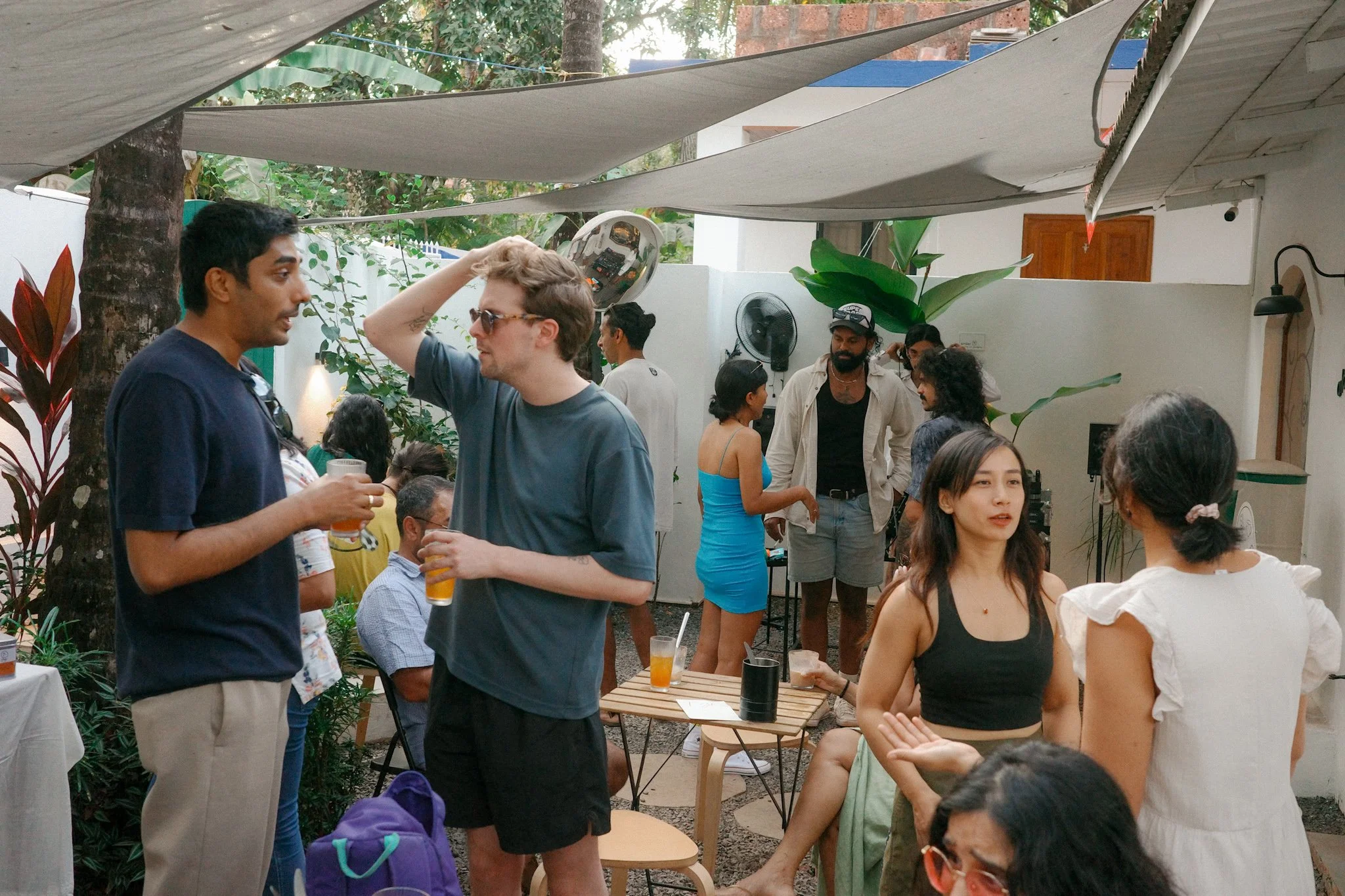 People socializing at an outdoor gathering with tropical plants, tables, and shade sails.