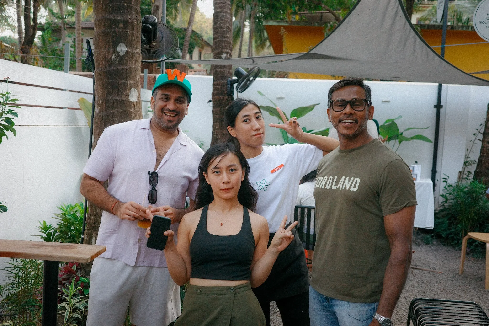Group of four young adults posing and smiling outdoors in a backyard, with trees and white fence in the background, during daytime.