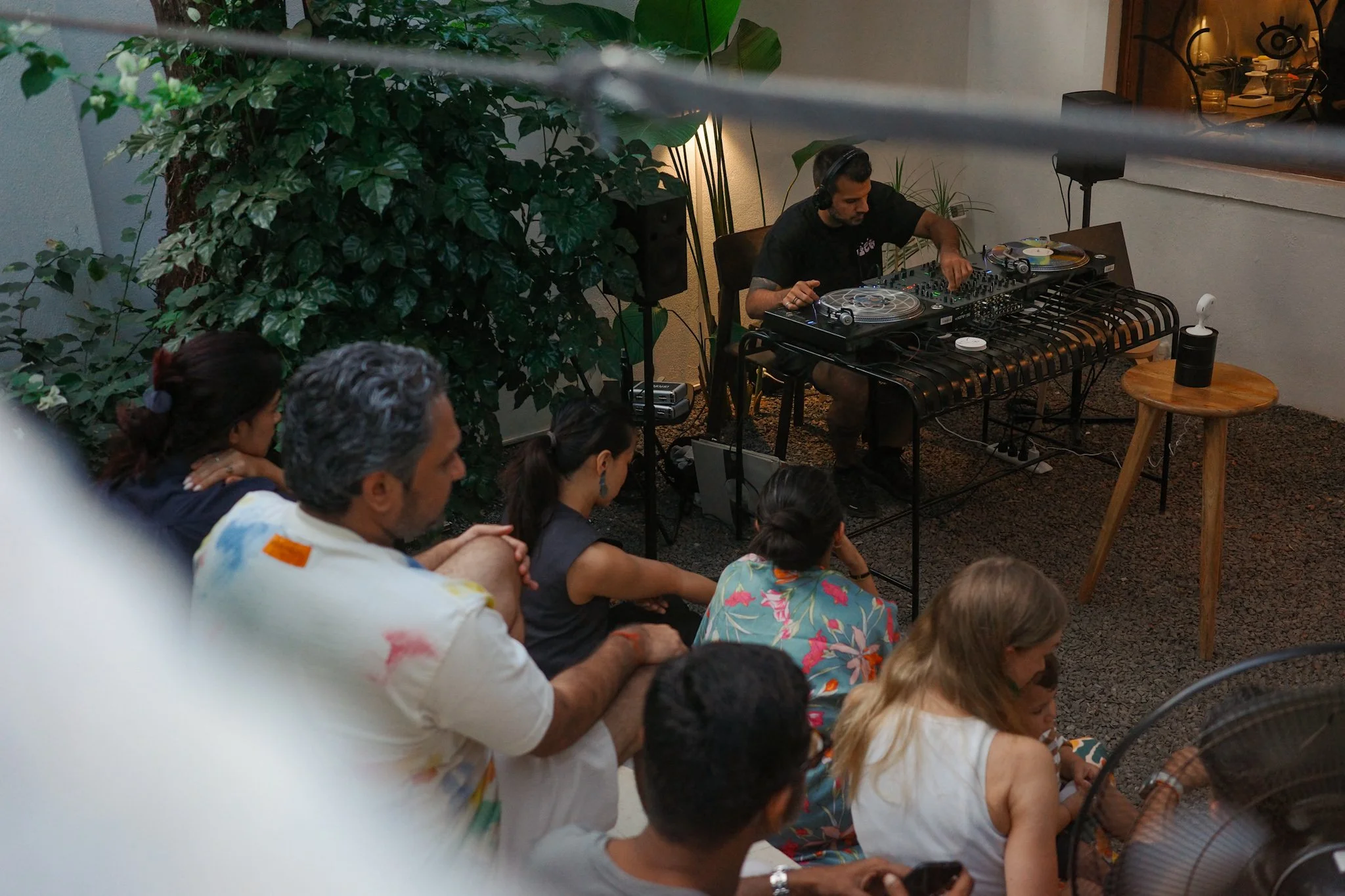 DJ performs music on a table with turntables and mixer, facing an audience seated on the floor indoors, with green plants and warm lighting in the background.