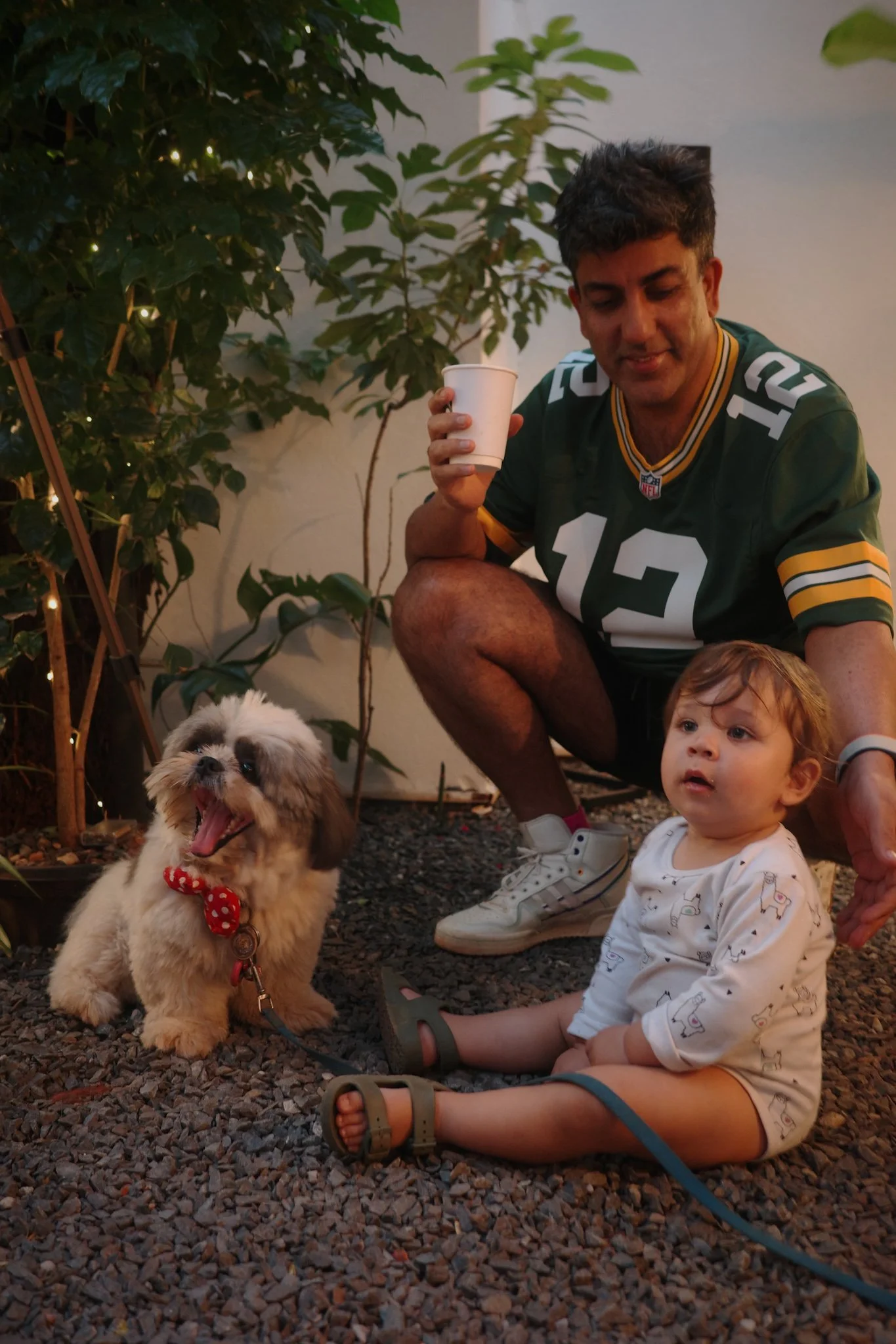 A man wearing a green football jersey with the number 12 on it, crouching next to a small child and a small fluffy dog. The man is holding a white cup, the child is sitting on gravel, and the dog is yawning with its mouth open, wearing a red polka-do