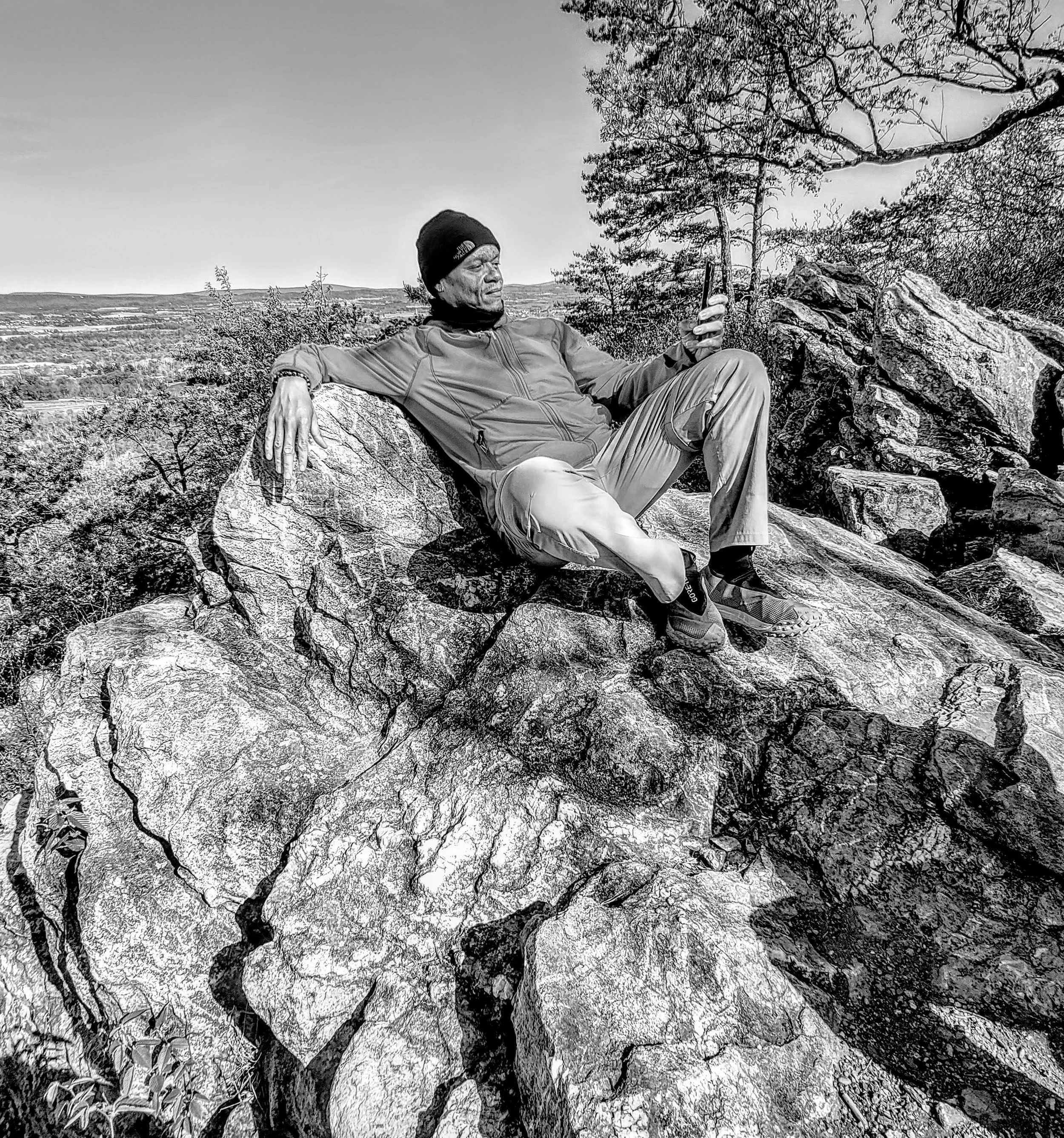 A man wearing a beanie, jacket, and hiking pants sits on a large rock in a natural outdoor setting with trees and a landscape in the background, looking at his phone.