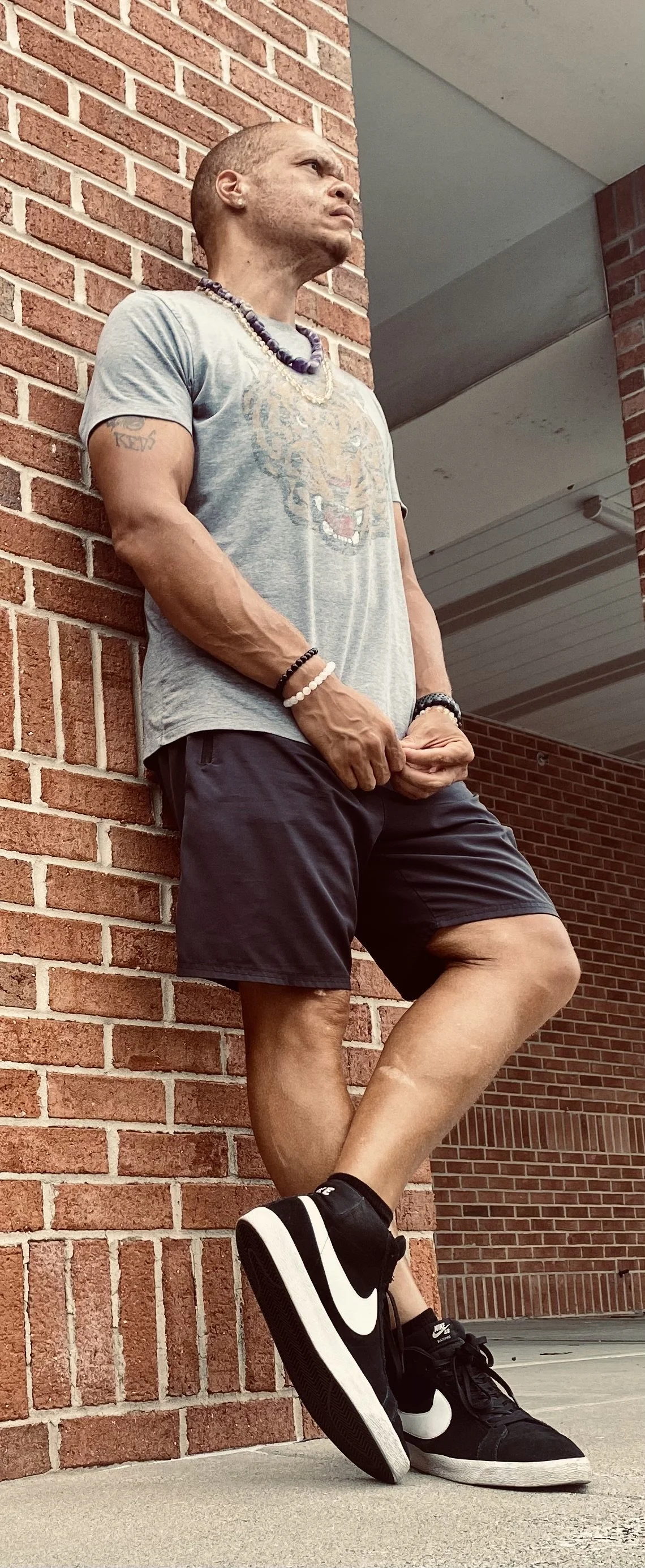 A man leaning against a brick wall outside, wearing a gray t-shirt, black shorts, and black Nike sneakers. He has muscular arms adorned with bracelets and a beaded necklace, and is looking off into the distance.