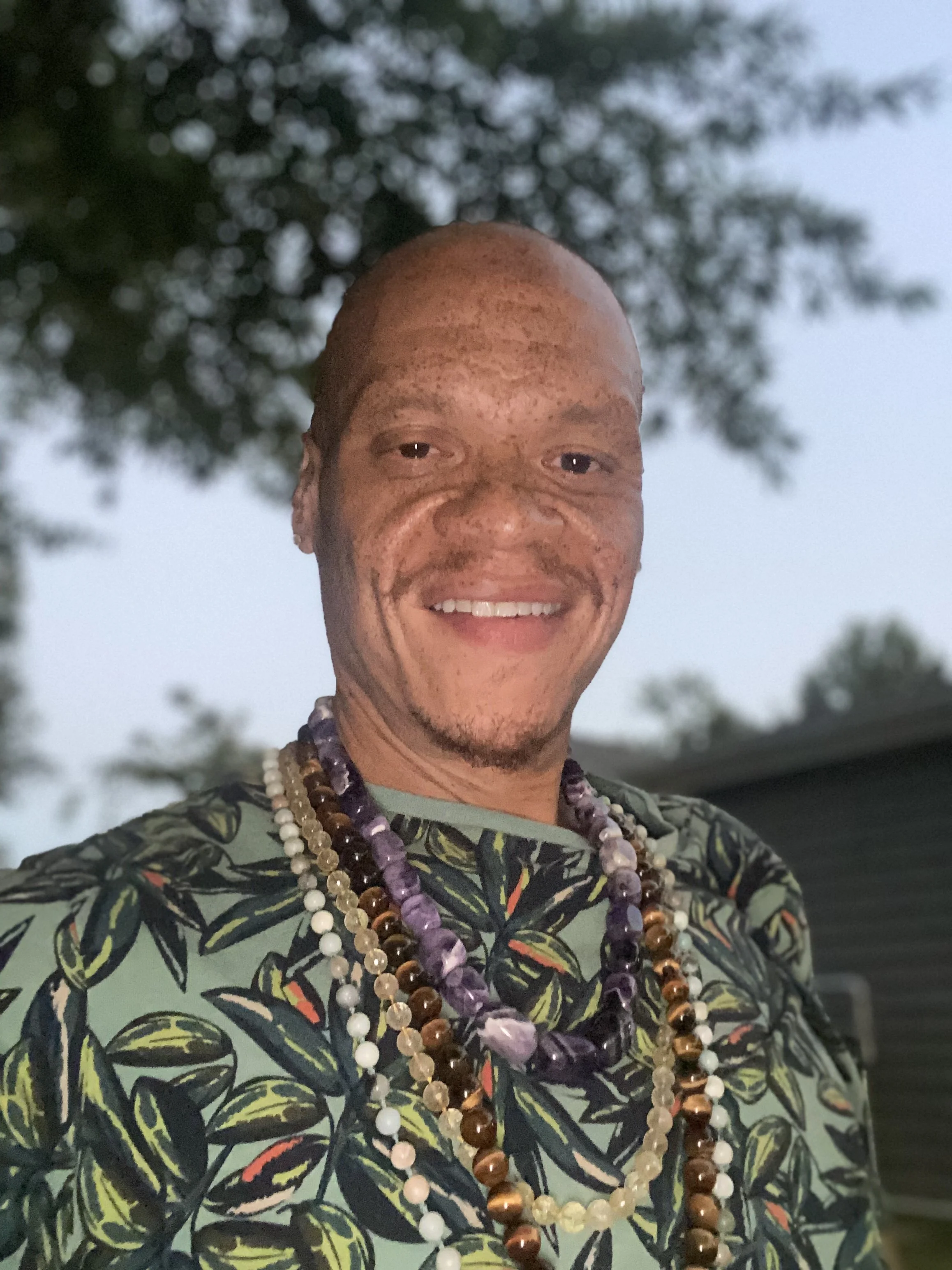 A man with a big smile, wearing a tropical-patterned shirt and multiple beaded necklaces, outdoors with trees and cloudy sky in the background.