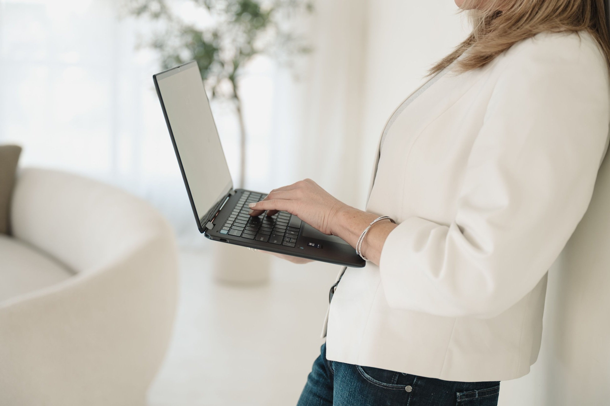 Woman in a white blazer holding a black laptop in a bright, modern room with white walls and furniture.