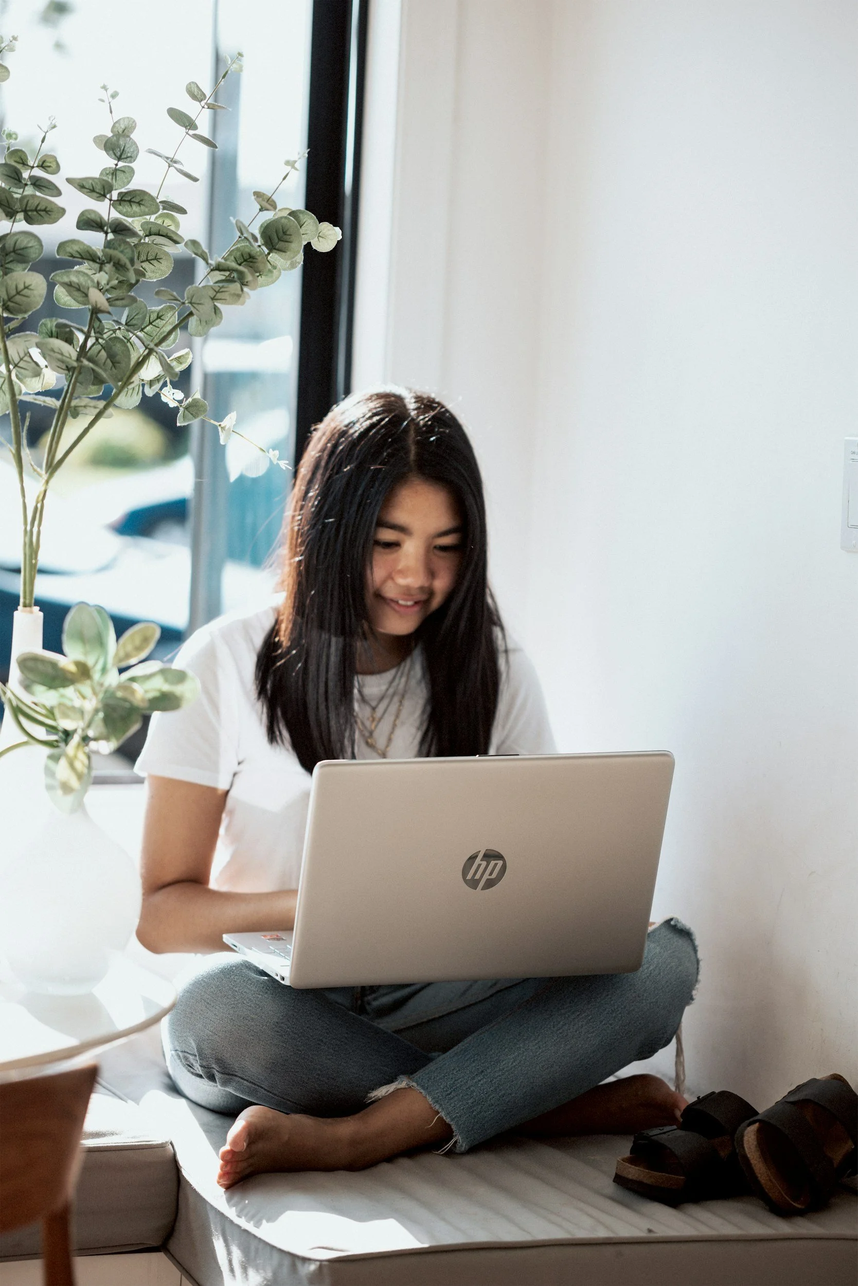 A young woman sitting cross-legged on a bed, working on a silver HP laptop, near a window with sunlight and a potted plant.