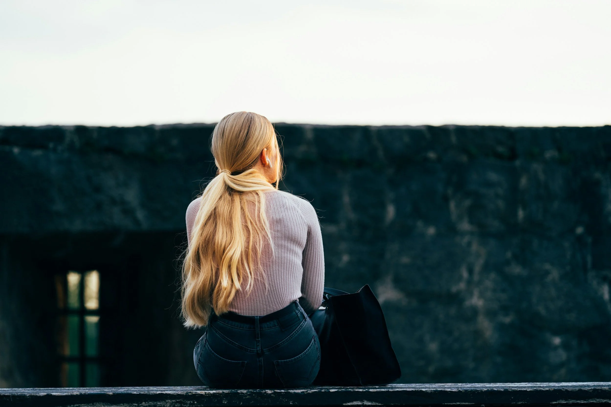 A woman with long blonde hair sitting outdoors on a ledge, looking away from the camera, with a dark stone wall in the background.