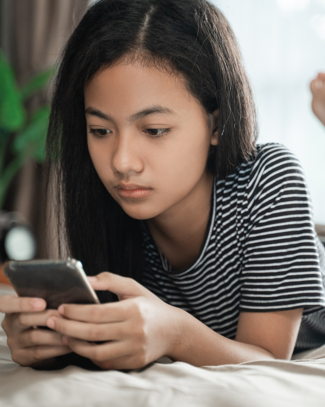 A young woman with straight black hair and light brown skin lying on her stomach and looking at her smartphone.