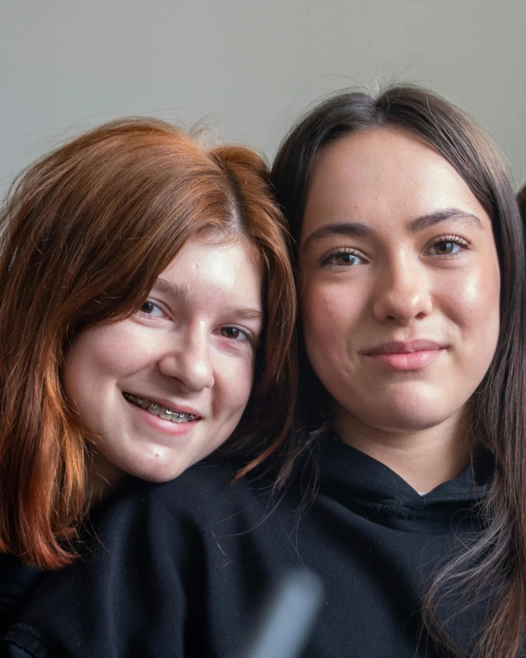 Two young women with light skin smiling closely together, one with red hair and braces, the other with dark brown hair.