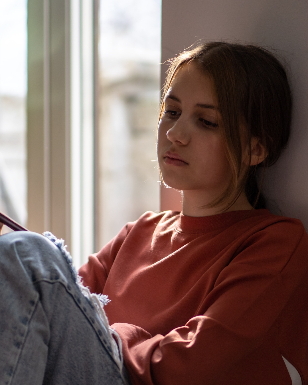 A young woman with short brown hair leaning against a wall near a window, looking down with a sad or reflective expression.