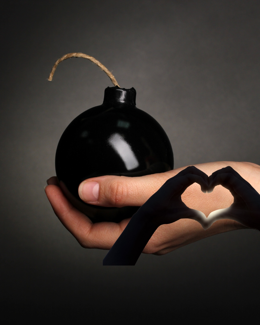 A hand holding a black spherical bomb with a lit fuse, with another hand making a heart shape in front of it.