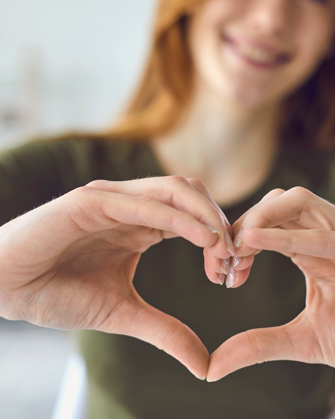 A woman with red hair smiling, making a heart shape with her hands.