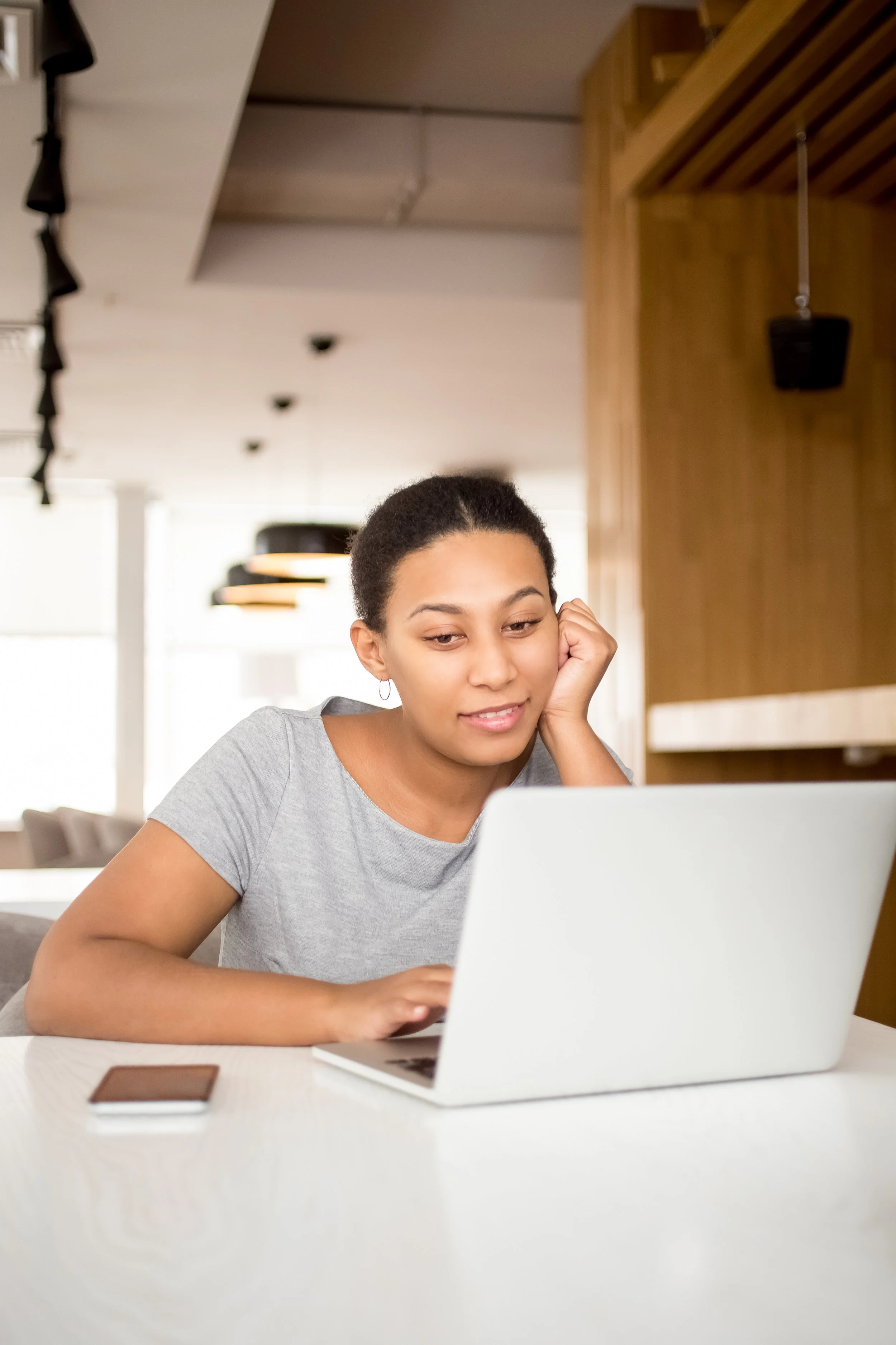 A young woman with short curly hair wearing a gray t-shirt, sitting at a white table, looking at a laptop with a slight smile, in a modern indoor setting.