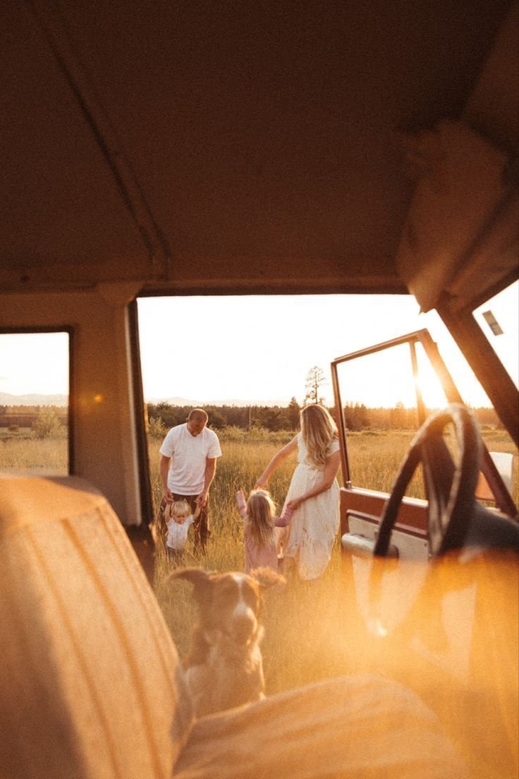 A family outside a vehicle during sunset in a grassy field. A woman and a man are holding two young girls' hands; a dog is nearby. The photo is taken from the vehicle's interior, overlooking the open door and part of the front seat.