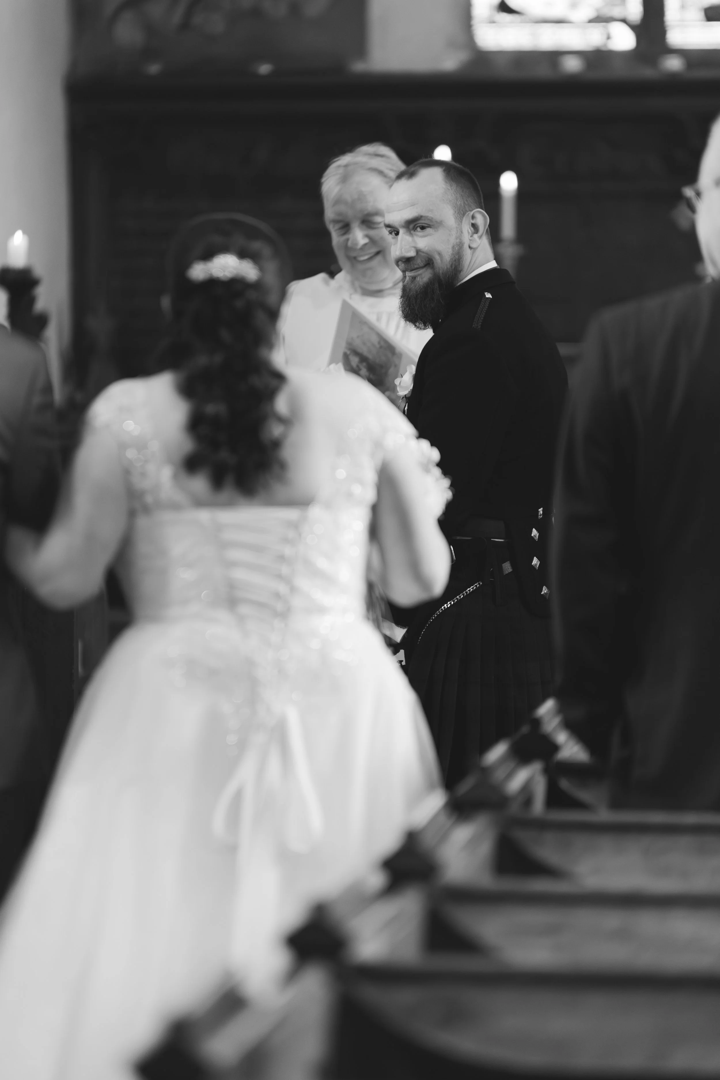Black and white photo of a wedding ceremony with a bride and groom standing before a minister, all surrounded by guests in a church setting.
