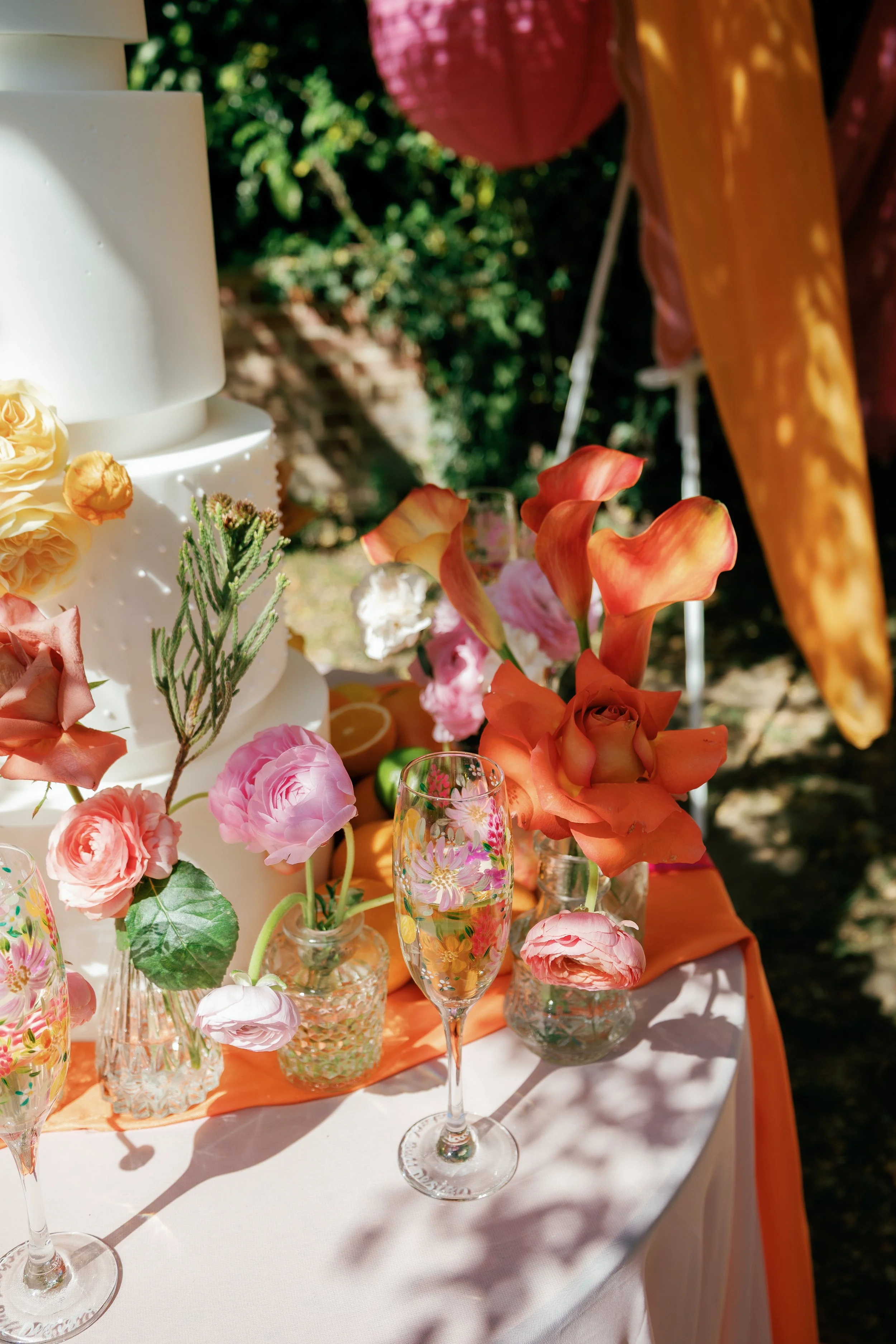 Decorative outdoor table with a white cake, pink and orange flowers in glass vases, two floral champagne flutes, and a pink paper lantern in the background.