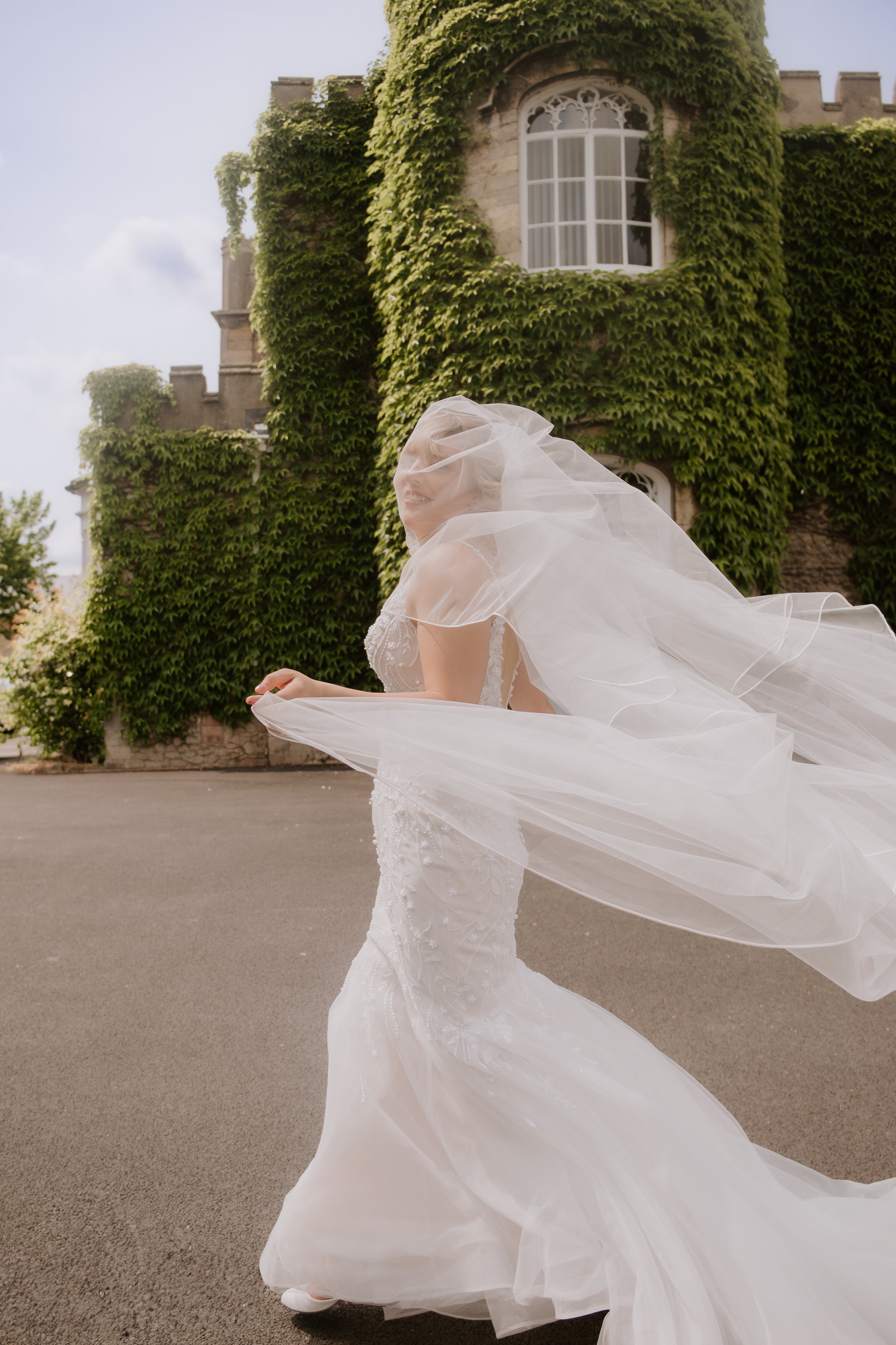 A bride in a white wedding gown with a long veil, smiling and twirling outdoors in front of a green ivy-covered building.