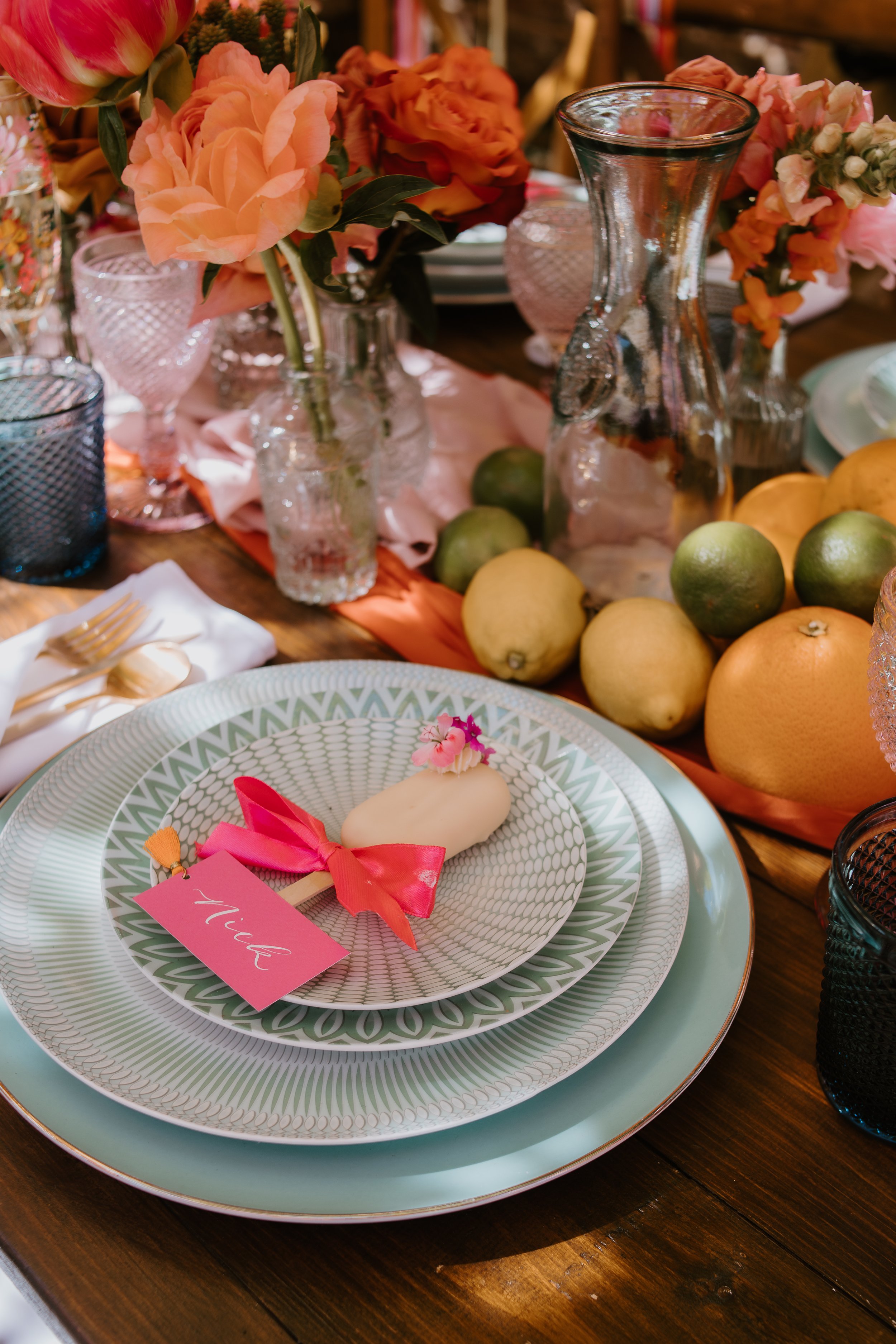 A table setting with floral arrangements, a juice pop, and a personalized place card reading 'Nina' surrounded by oranges and lemons.
